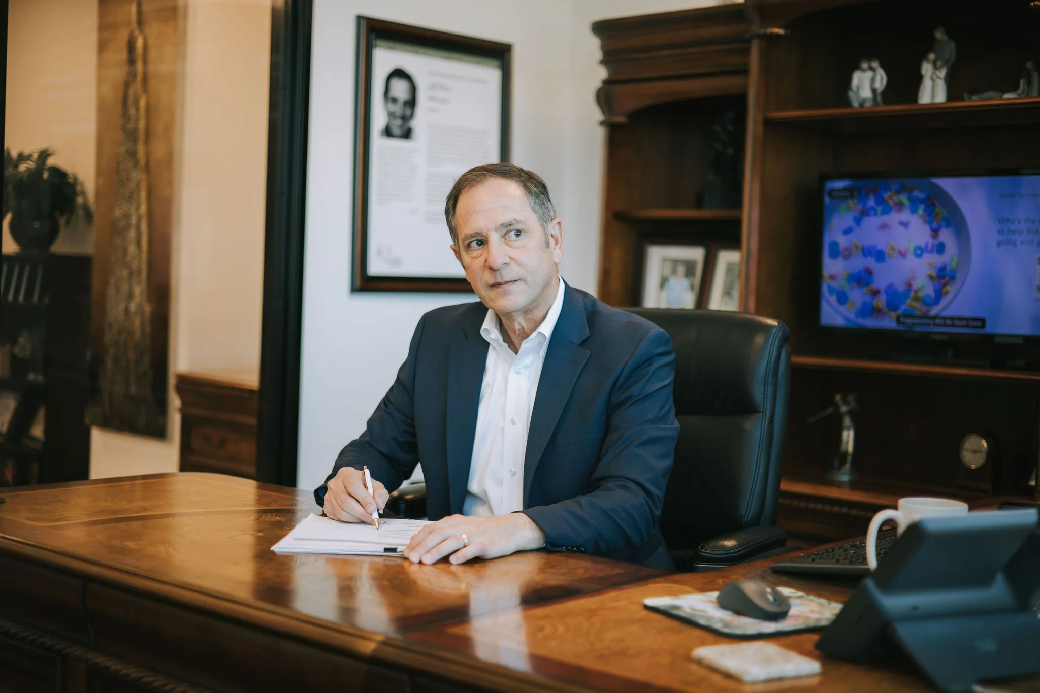 Jeff Klein of Klein Investment Strategies, financial investment firm located in Bend, OR holding a pen over a stack of papers, in an office with wooden shelves, framed photos, and a framed portrait in the background.