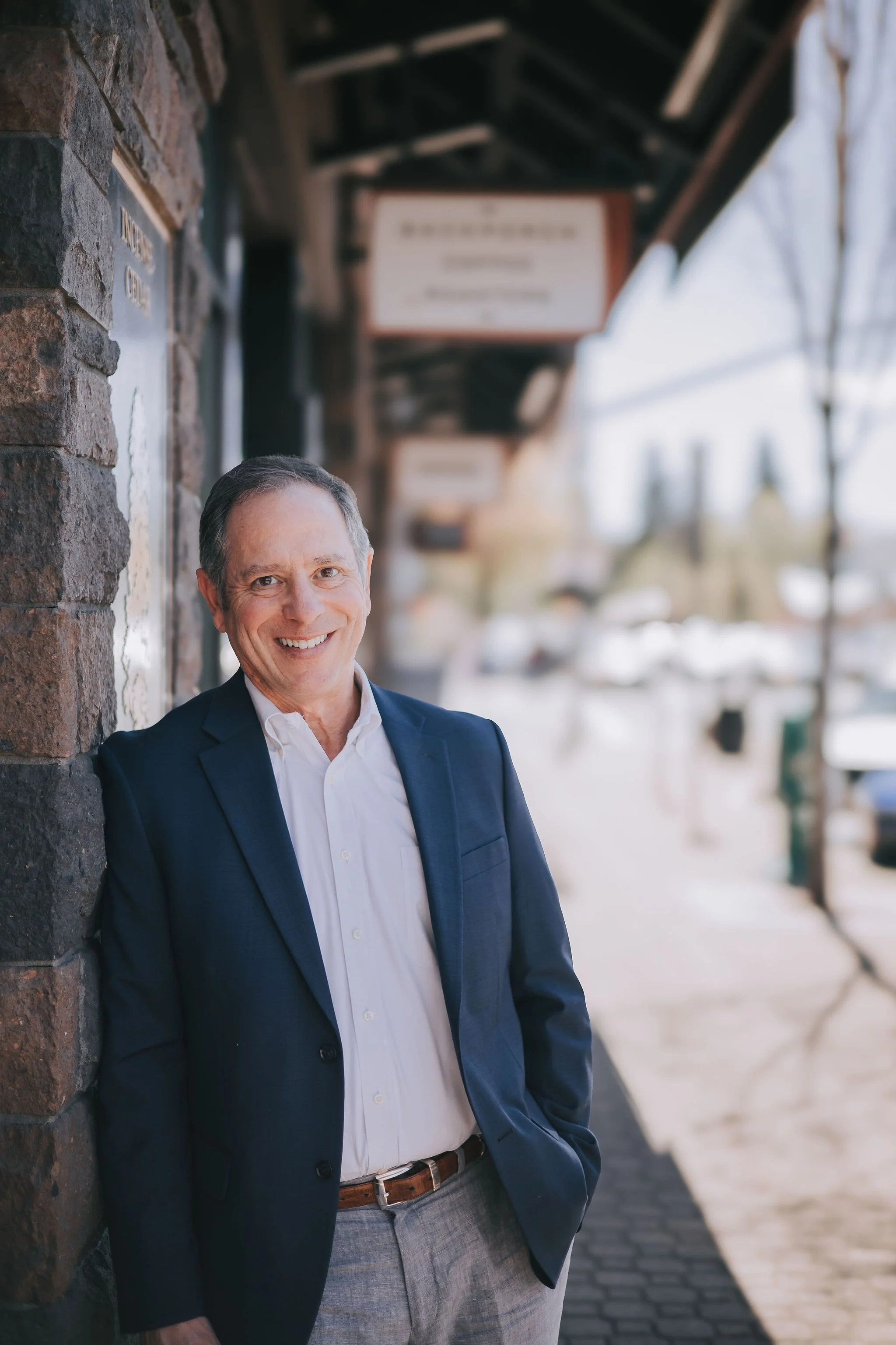 A smiling middle-aged man in a dark blue blazer, white shirt, and gray pants standing against a brick wall on a city sidewalk, with blurred buildings and trees in the background.