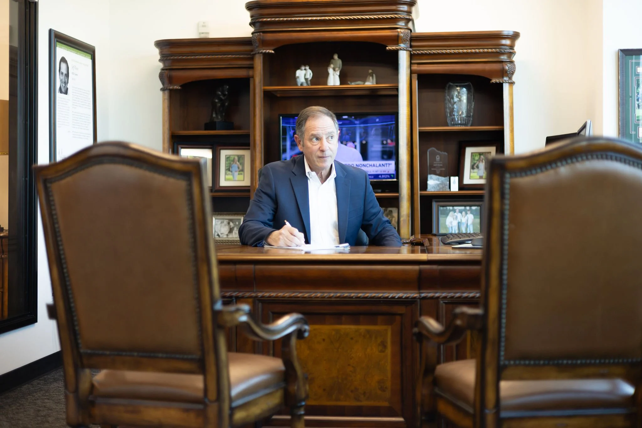 Jeff Klein of Klein Investment Strategies, financial investment firm located in Bend, OR. Office with wooden shelves, framed photos, and a television in the background. The photo is taken through a gap between two chairs.