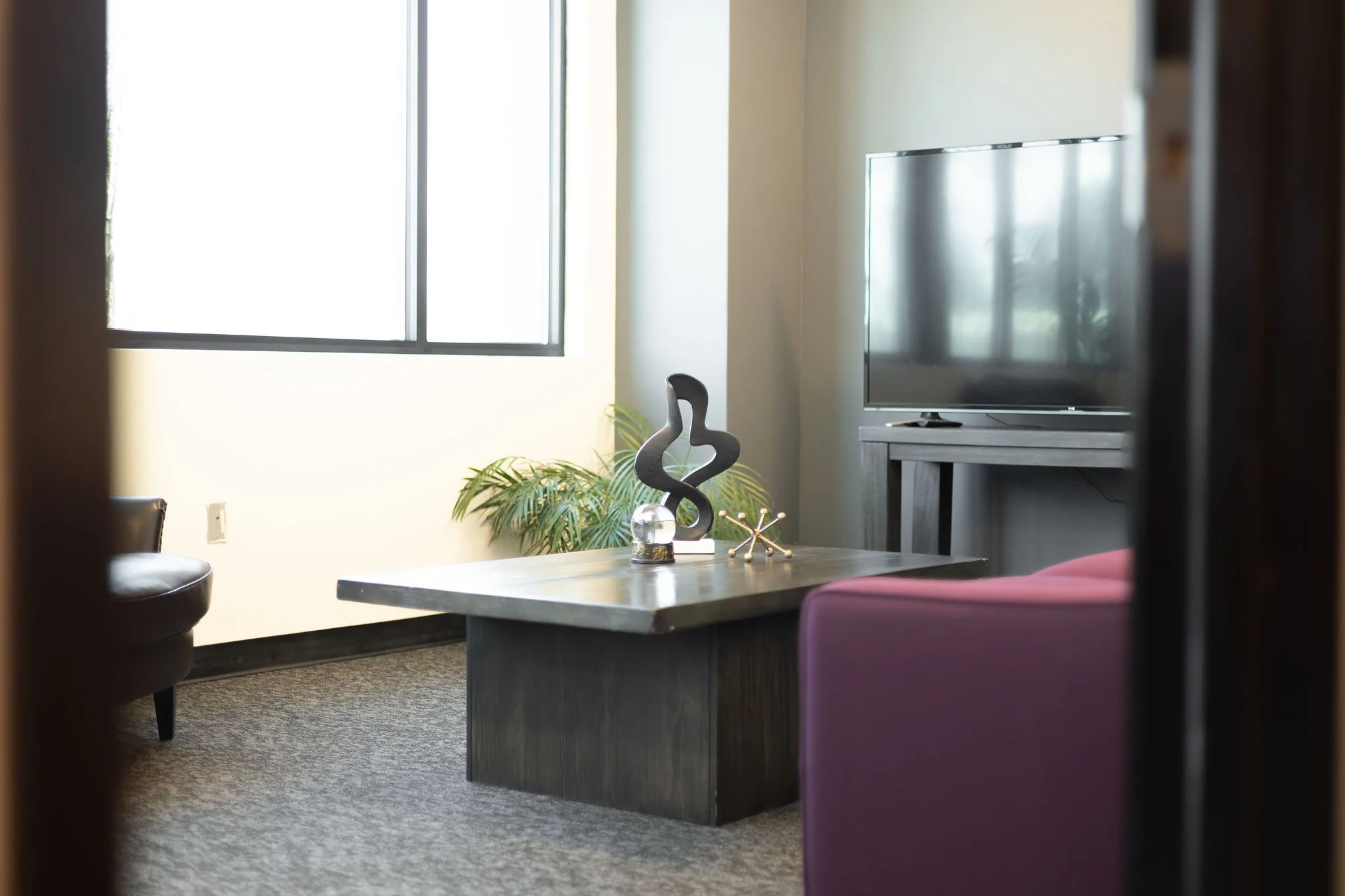 Living room with a large window, flat-screen TV on a stand, decorative sculpture and plant on a coffee table, purple armchair, and a black chair.