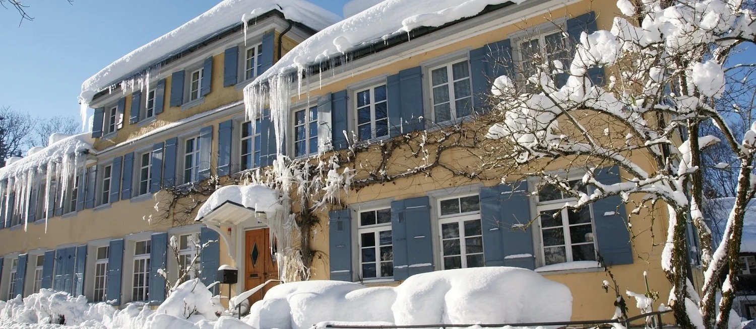Mehrstöckiges Haus mit blauen Fensterläden, umgeben von schneebedeckten Bäumen und Hügeln, Eiszapfen hängen von dem Dach, das im Winter von Schnee bedeckt ist.