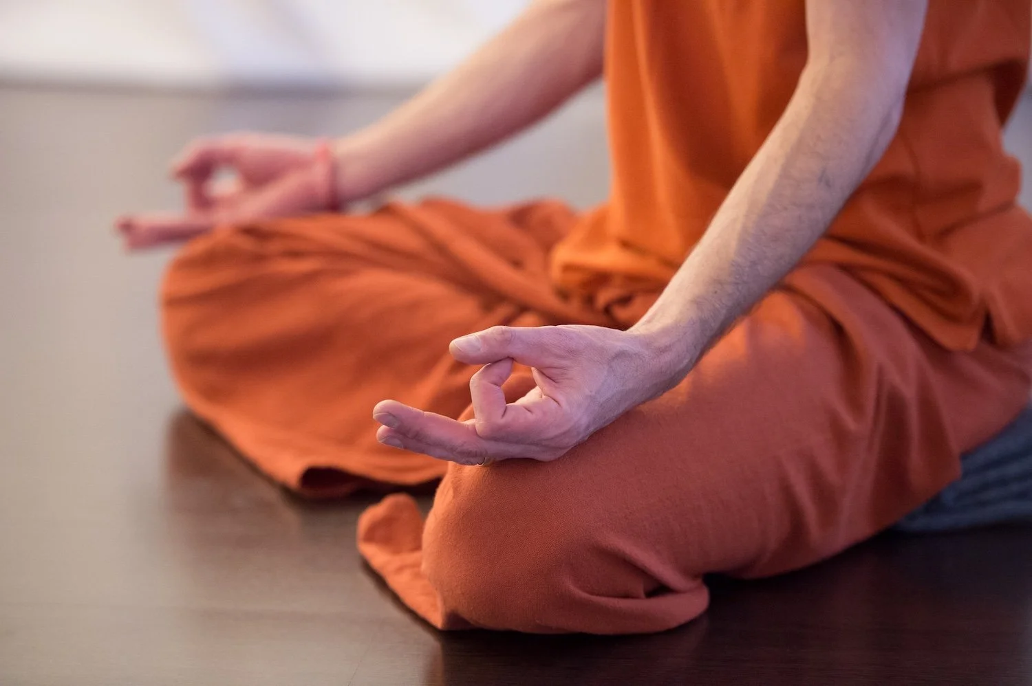 Eine Person sitzt im Schneidersitz auf dem Boden, trägt eine orange Kleidung und übt eine Meditationshaltung mit der Hand in der Mudra-Position.