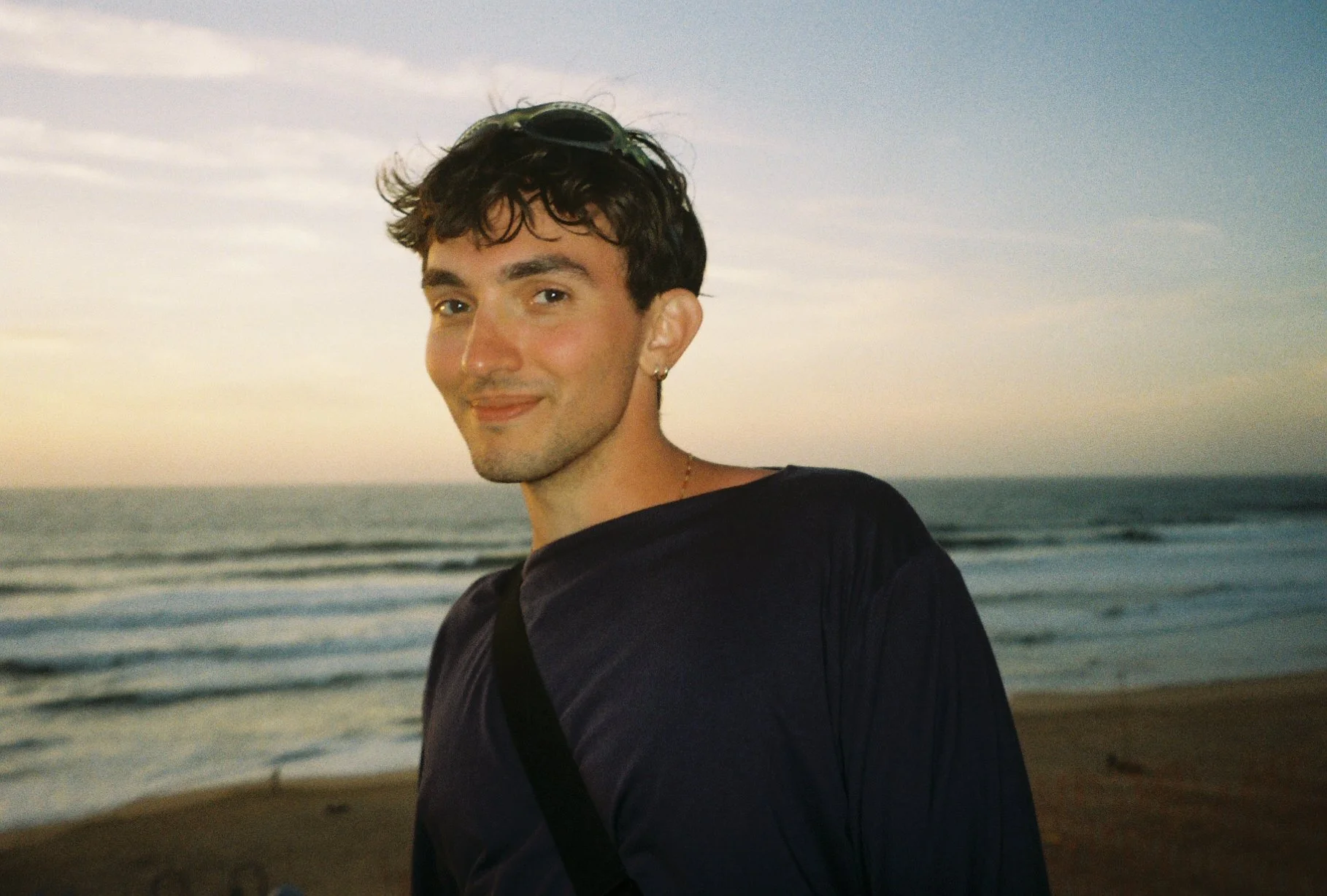 A young man with curly dark hair, wearing sunglasses on his head, earrings, and a dark shirt, standing on a beach at sunset with the ocean and sky in the background.