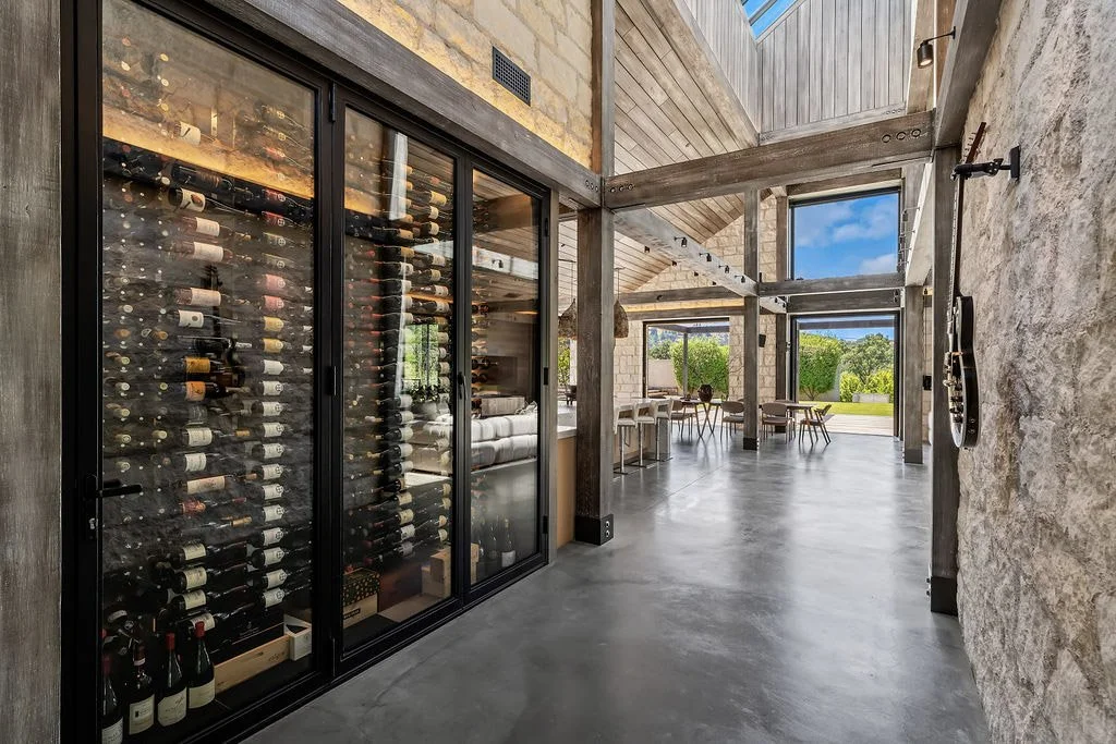 Wine bottles stored inside a glass-front wine fridge in a modern, rustic-style room with stone and wood walls, concrete floor, large windows, and outdoor seating visible outside.