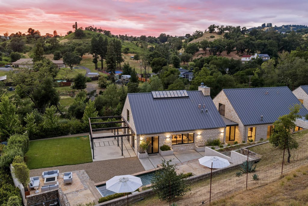 Aerial view of modern house with stone exterior, metal roof, and backyard with patio, pool, and umbrellas, surrounded by trees and green rolling hills during sunset.
