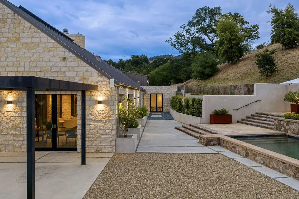 Exterior view of a modern house with stone walls, outdoor lighting, and a patio area, featuring a small pool and landscaped steps.
