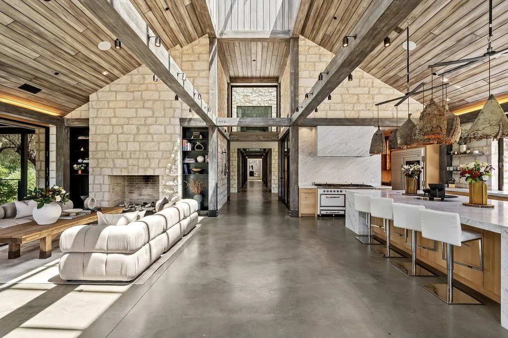 Open-concept living room and kitchen with vaulted wooden ceiling, stone and wood accents, a plush beige sofa, white bar stools, and rustic light fixtures.
