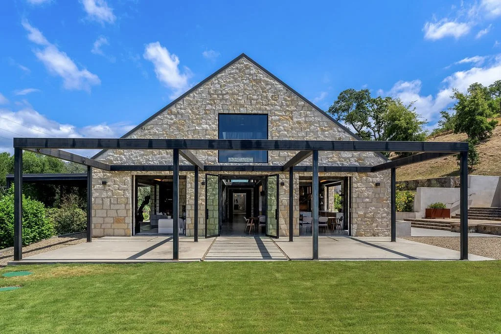 Modern house with stone exterior and large glass doors, black metal framework, and a manicured green lawn under a partly cloudy sky.