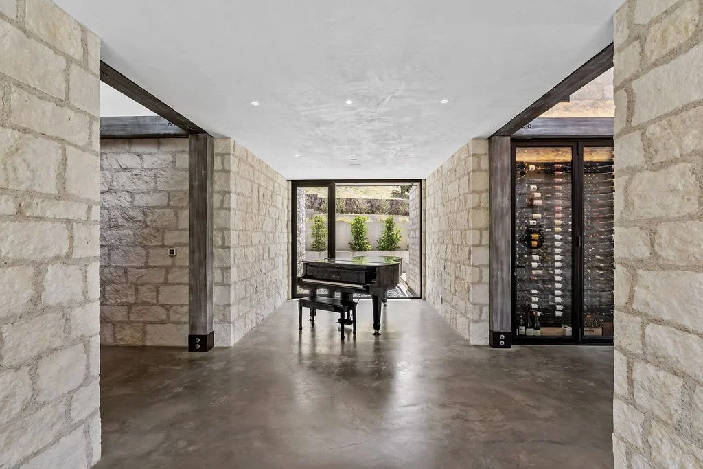Modern interior with a black grand piano in the center, stone walls, a large glass door leading outside with greenery, and a wine fridge on the right.