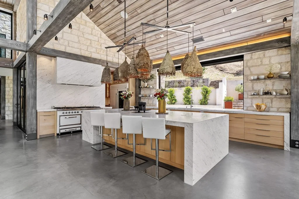 Modern kitchen with a large white marble island, white barstools, wooden cabinets, and decorative pendant lights made of rustic materials. Large window with a view of greenery outside.