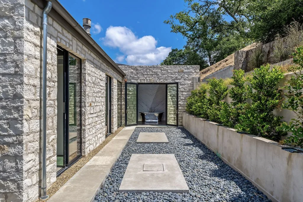 A modern outdoor patio with stone walls, gravel ground, and stepping stones, enclosed by a concrete wall with greenery, leading to a glass door with black frames and a window in a brick building.