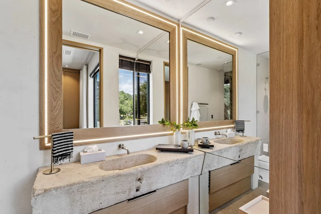 Modern bathroom with double vanity, concrete sinks, large mirrors with wood frames, and a window showing trees outside.