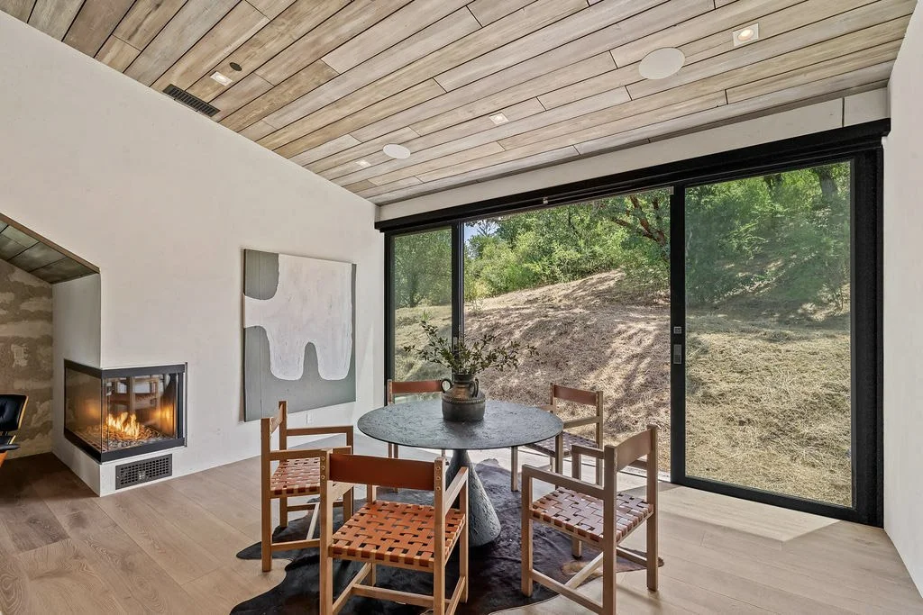 Modern dining room with a round table and four wooden chairs, large sliding glass doors showing an outdoor hillside, a corner fireplace, and a wooden ceiling.