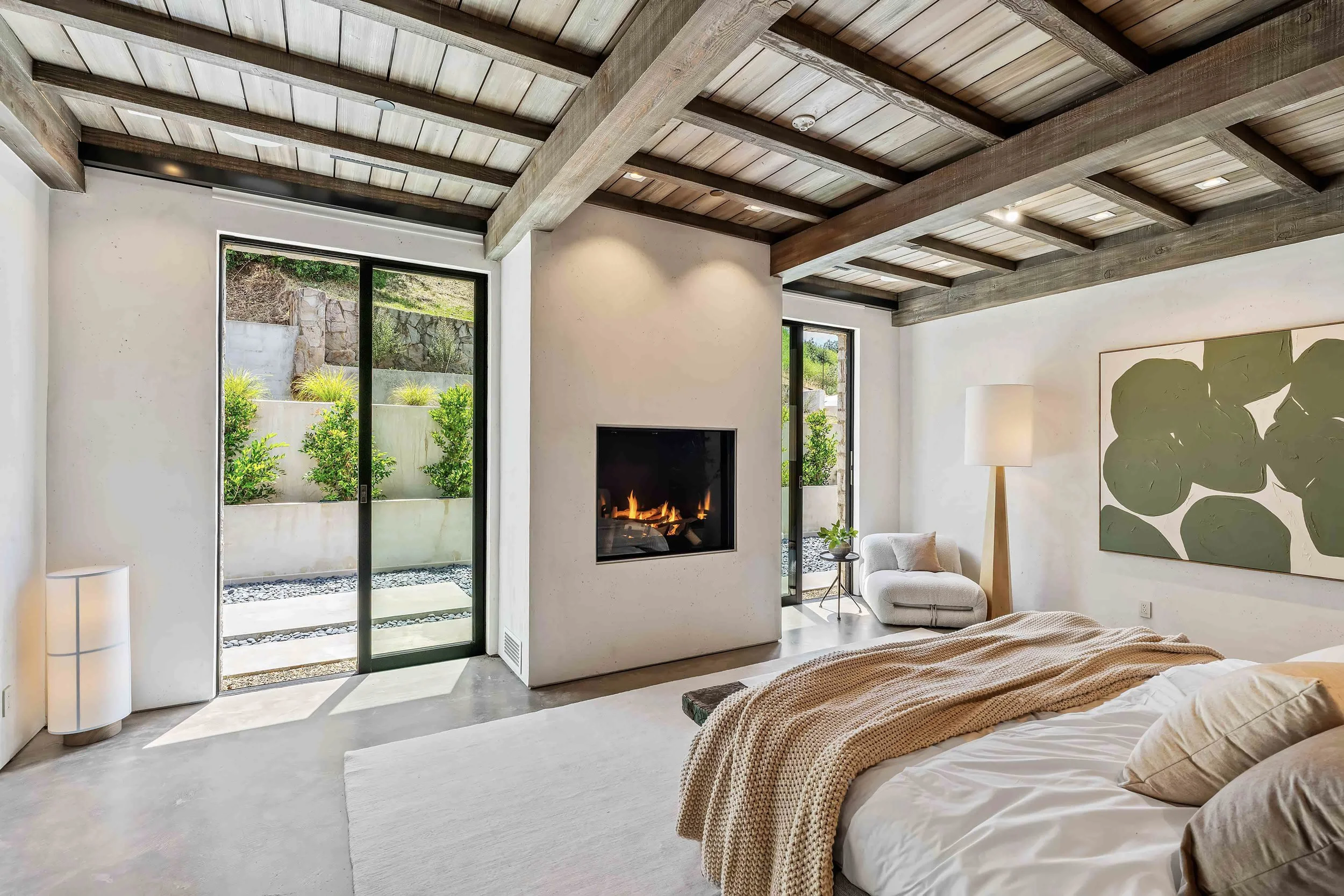Modern bedroom with a wood-beam ceiling, a large window showing outdoor greenery, a fireplace, a side table with a small plant, and an abstract green artwork on the wall.