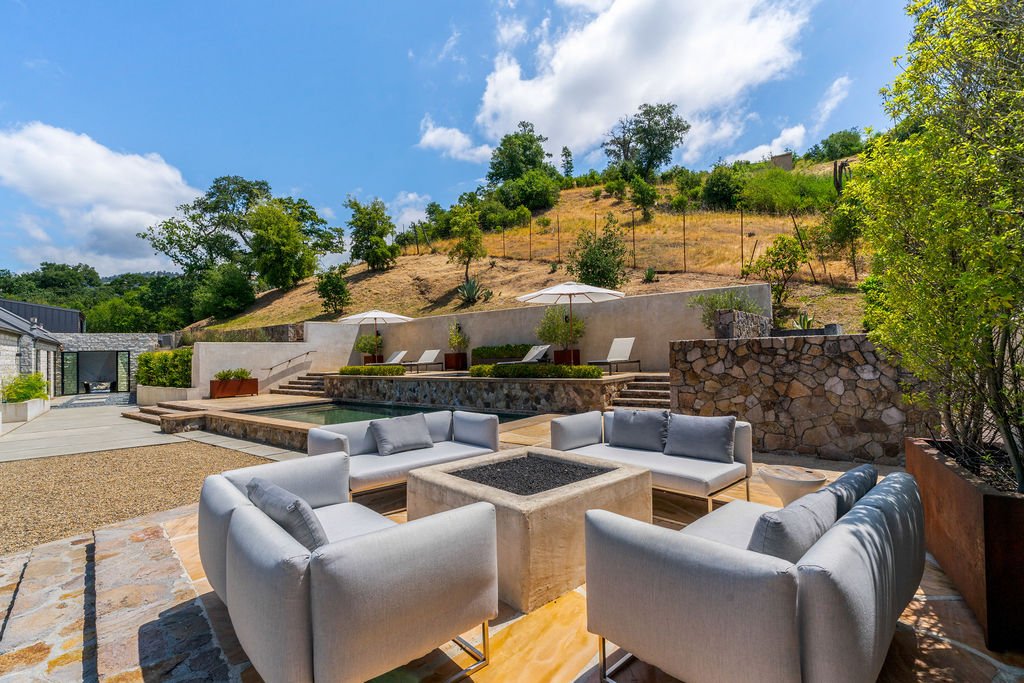 Outdoor patio with modern white seating around a fire pit, overlooking a small pool, with lounge chairs, umbrellas, and hillside in the background under a partly cloudy sky.