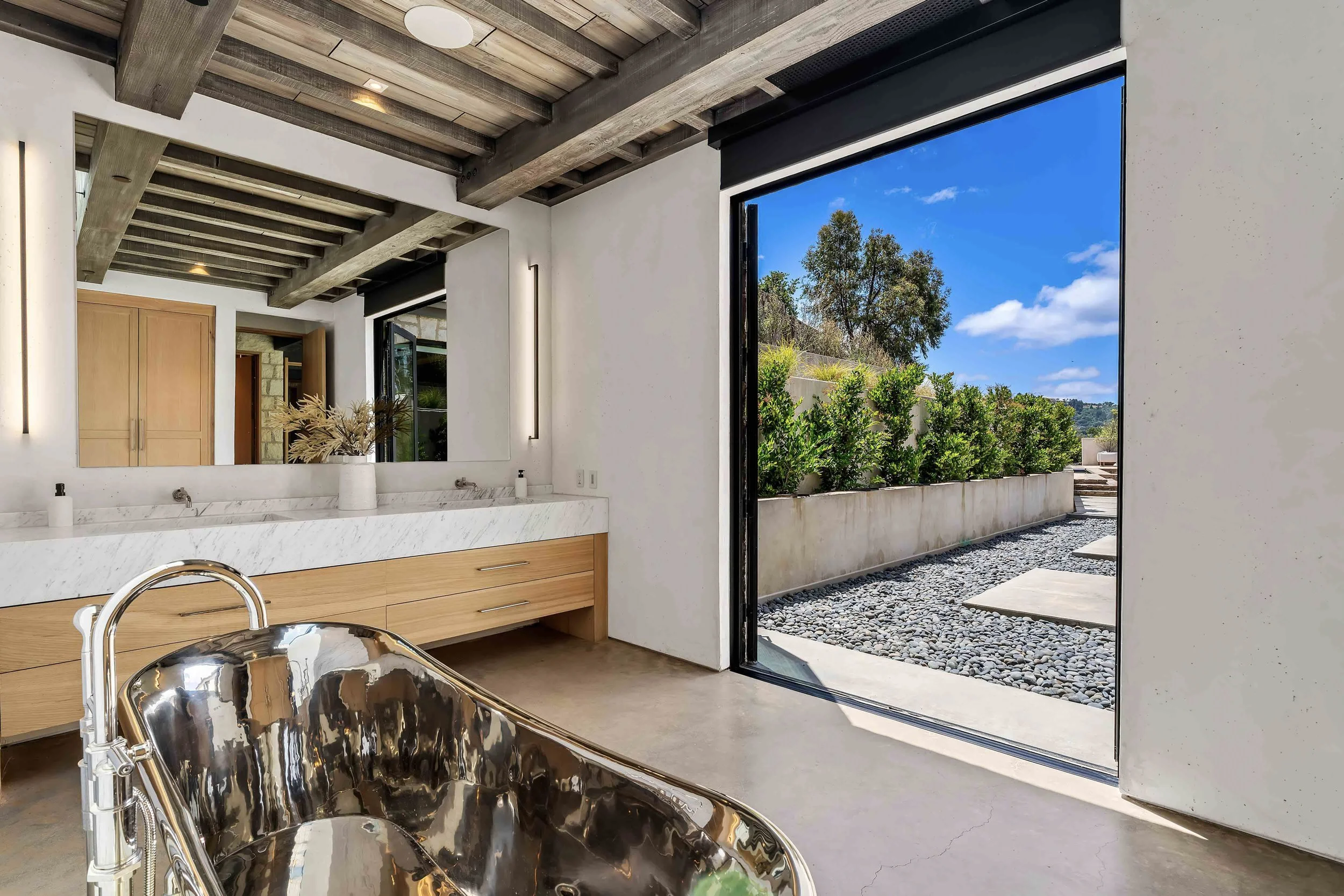 Modern bathroom with a large mirror, wooden cabinets, and a stainless steel bathtub open to an outdoor patio with greenery and a blue sky.