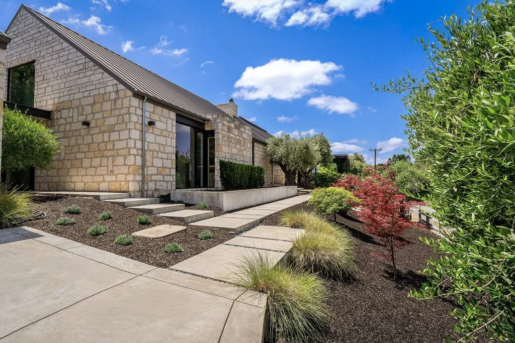 Modern house with stone exterior, large windows, and landscaped garden with trees, bushes, and a paved walkway.