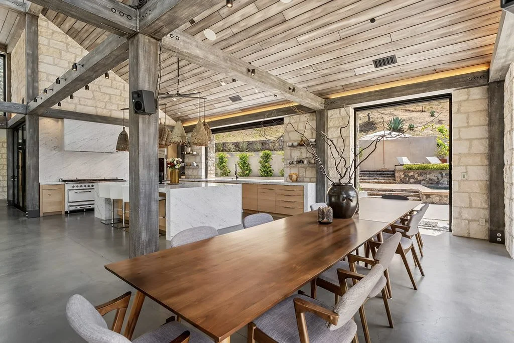 Modern open-concept kitchen and dining area with a large wooden dining table, surrounded by beige and gray chairs, featuring a black vase with branches as a centerpiece, in a room with wooden ceiling, stone and wood walls, and large sliding door lead