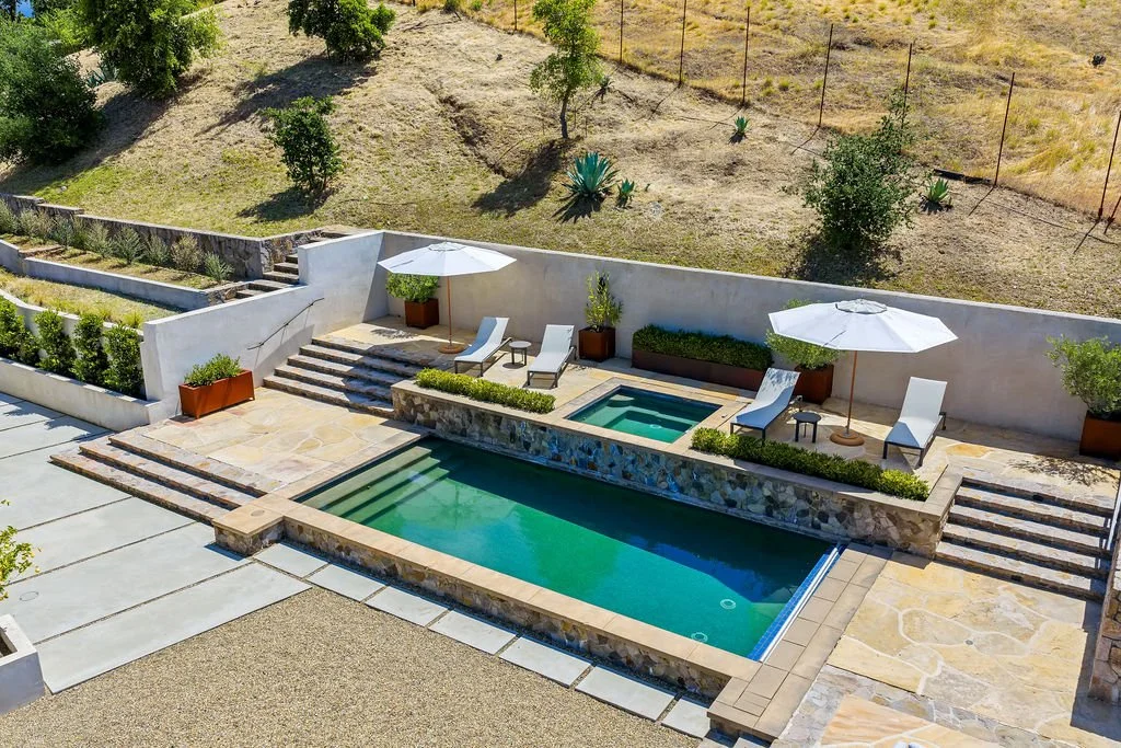 A backyard with a swimming pool and hot tub, surrounded by a patio with lounge chairs, umbrellas, and potted plants, overlooking a hill with grass, trees, and desert plants.