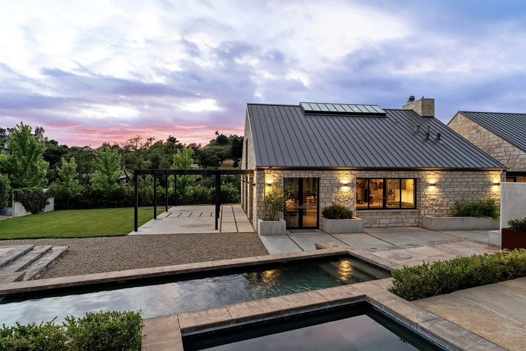 Modern house with stone exterior, metal roof, and large windows, surrounded by a landscaped yard with a swimming pool, trees, and outdoor lighting at sunset.