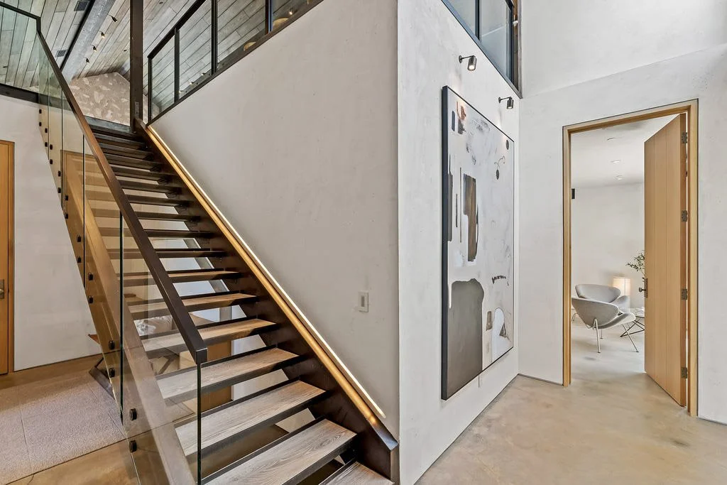 Modern interior of a house showing a wooden staircase with glass railing, abstract wall art, and a doorway leading to a living room with a white chair and a floor lamp.