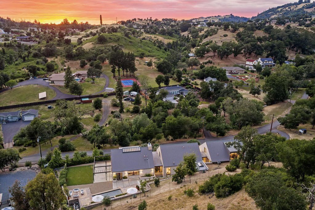 An aerial view of a residential neighborhood on hills at sunset, featuring houses surrounded by trees, winding roads, tennis courts, and a mountainous landscape.