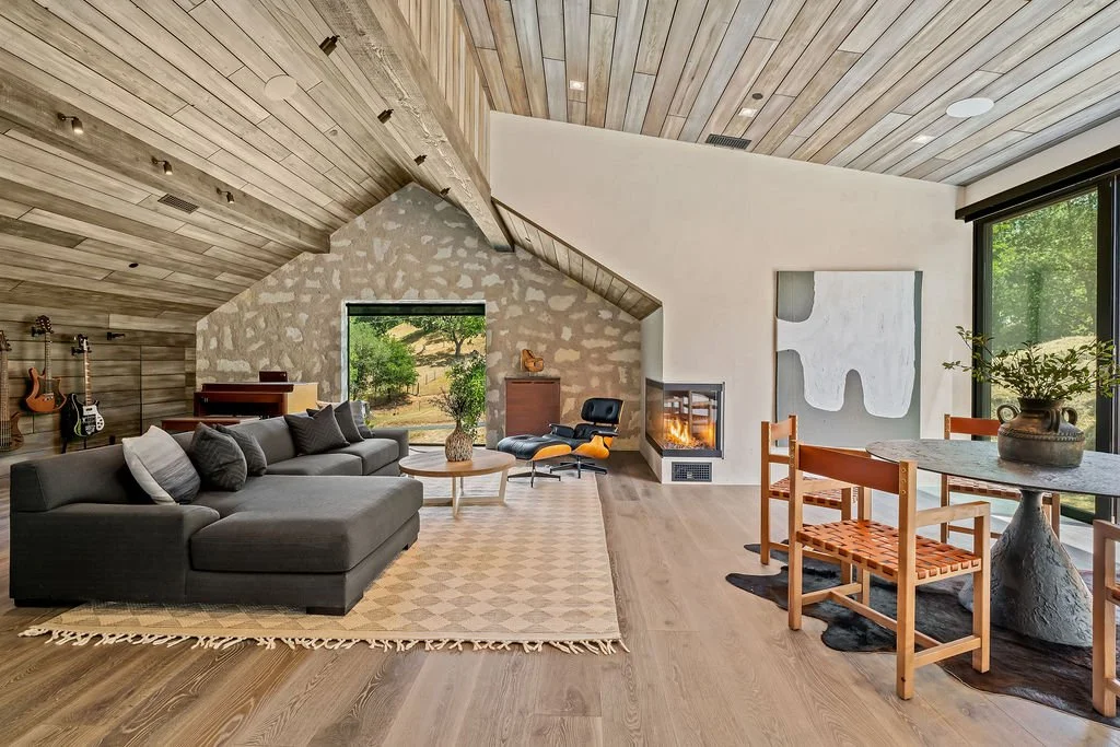 Modern living room with a gray sectional sofa, a round coffee table, and a fireplace. Guitar instruments are mounted on the wall, and an abstract painting hangs on the white wall. A wooden dining table with chairs is near large sliding glass doors, with natural light and greenery outside.