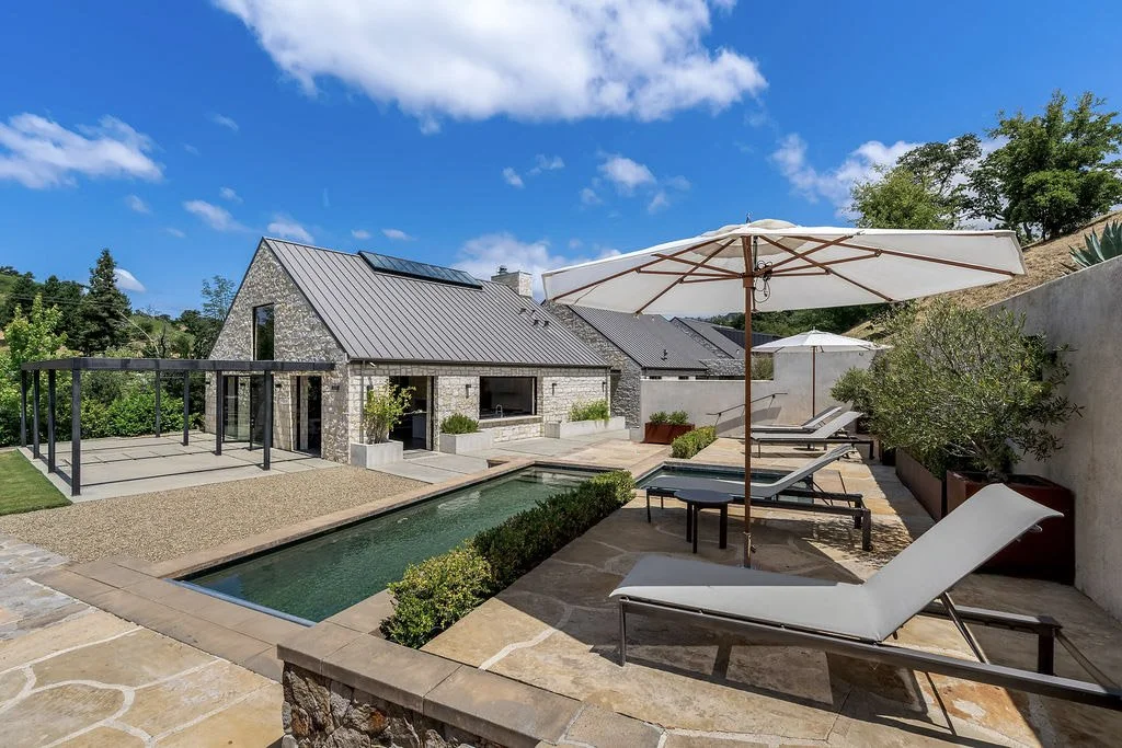 Backyard with a swimming pool, lounge chairs with umbrellas, and a modern house with solar panels on the roof under a partly cloudy sky.