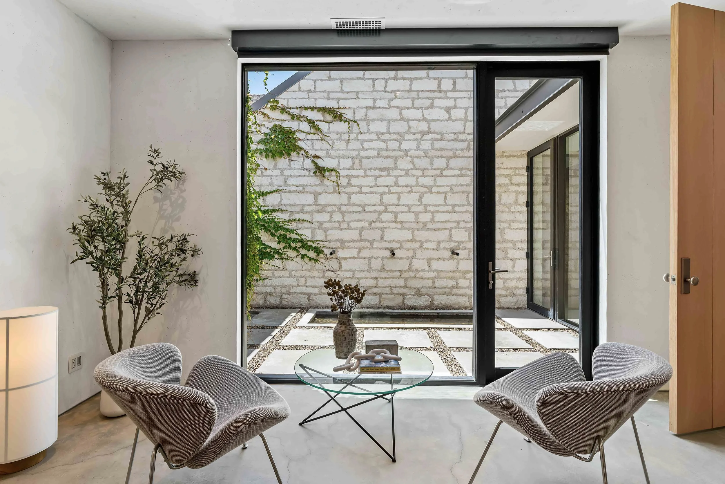 Modern interior living space with four beige upholstered chairs arranged around a glass coffee table, large sliding glass door leading to an outdoor patio with a brick wall, potted plants, and geometric paving stones.