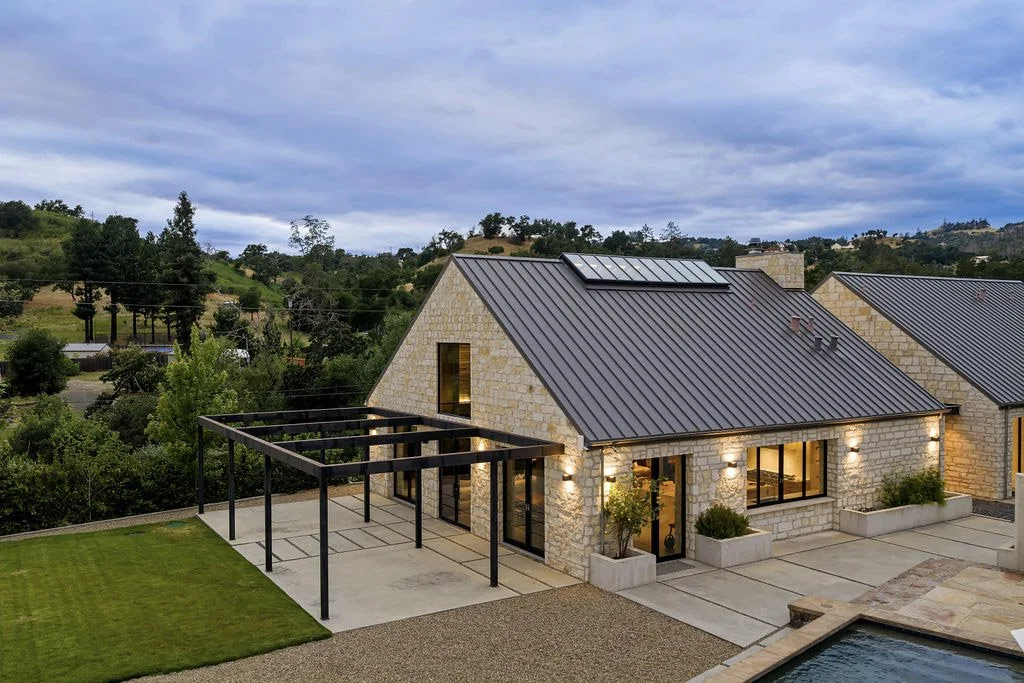 Modern house with stone exterior, metal roof, and large illuminated windows, situated in a hilly landscape with trees and a cloudy sky.