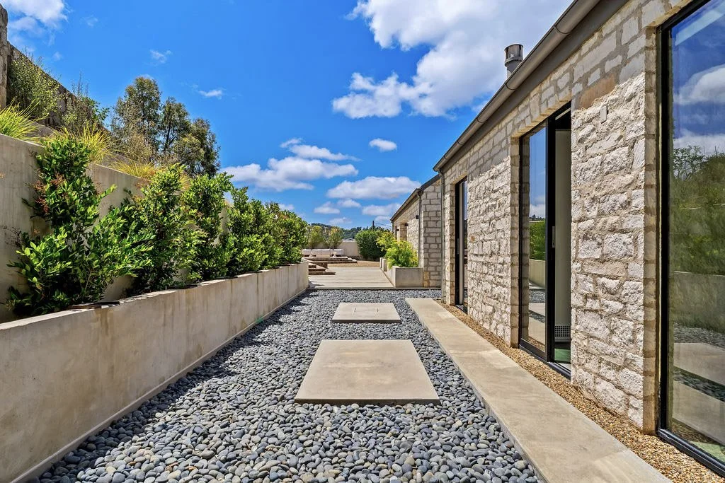 Modern house exterior with stone walls, large glass sliding doors, concrete stepping stones, and a gravel walkway, with a landscaped garden on the left side and a blue sky with clouds overhead.