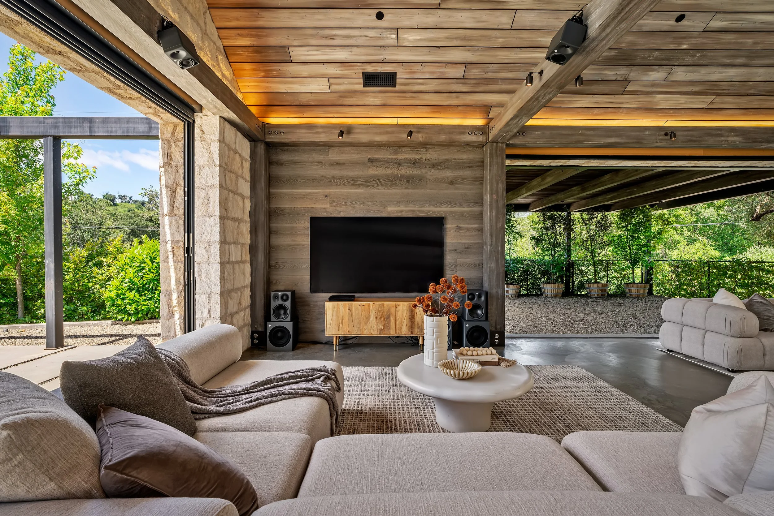 A modern living room with large beige sectional sofas, a round white coffee table, and a wooden media console with a flat screen TV and speakers. The room has an open view of lush greenery and trees outside, with natural light, wooden ceiling beams, 