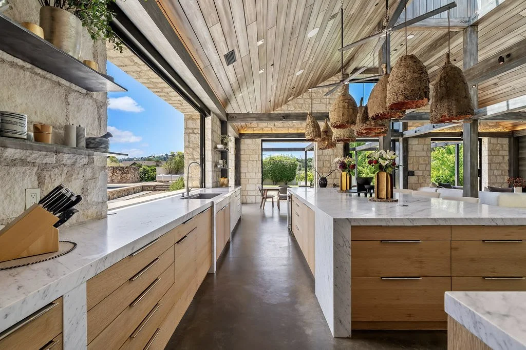 Modern kitchen with marble countertops, wooden cabinets, open view, hanging woven light fixtures, and lush greenery outside.