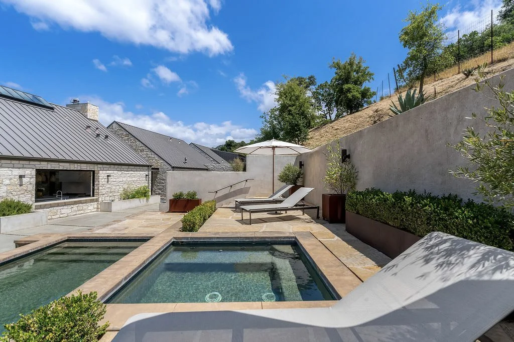 Backyard with a small rectangular pool, two lounge chairs with an umbrella, potted plants, and a stone house with a metal roof under a blue sky with clouds.