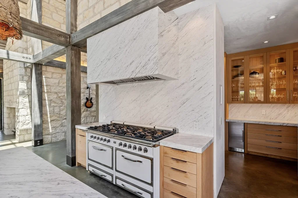 A modern kitchen with a white marble backsplash and countertops, a vintage white stove, wooden cabinets, and a glass-front display cabinet filled with glassware, with exposed stone walls and wooden beams.