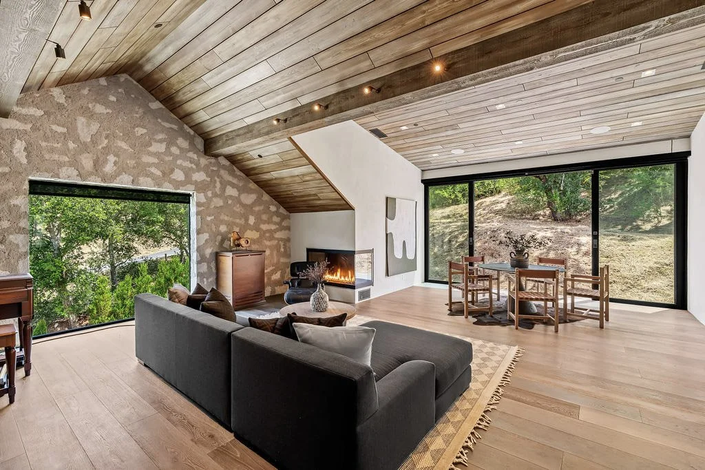 Modern living room with wood-paneled ceiling, stone accent wall, fireplace, large windows showing outdoor greenery, and minimalistic furniture.