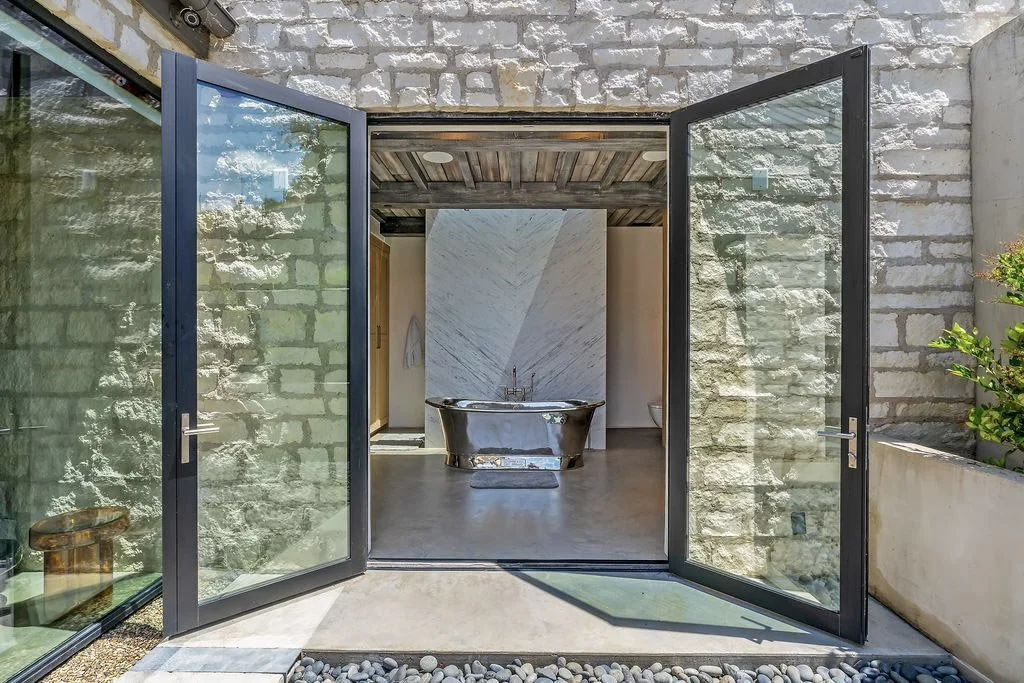 Open glass doors leading to a modern bathroom with a freestanding bathtub and a marble wall.