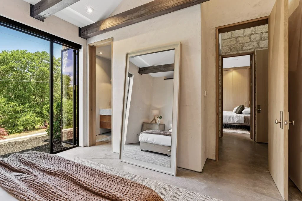 Interior of a bedroom with a large mirror leaning against the wall, a bed with a blanket, and a sliding glass door that opens to a green outdoor view.