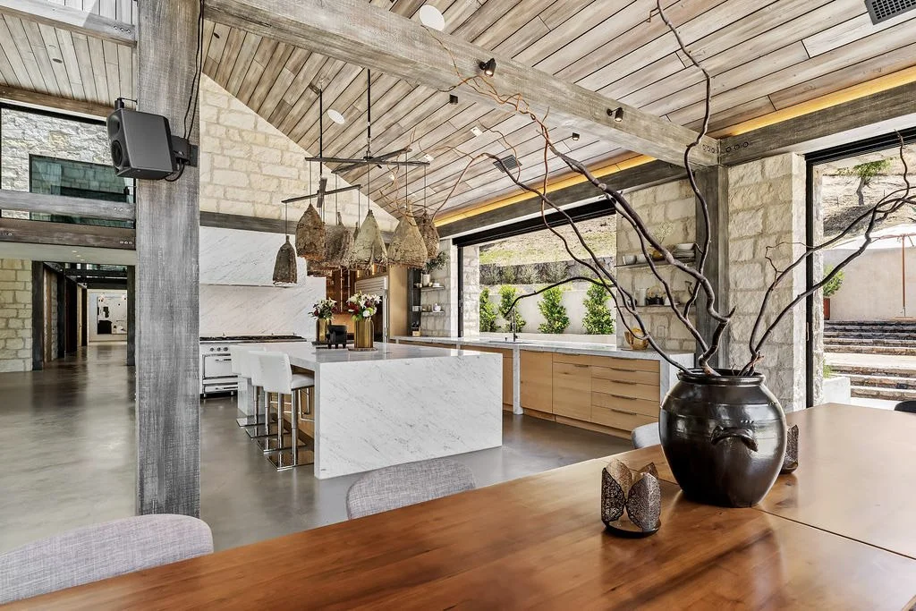 Contemporary open-concept kitchen and dining area with wooden and marble accents, large windows, and decorative branch arrangement in a black vase.