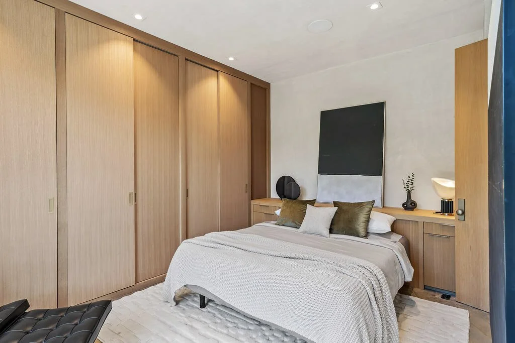 A modern bedroom with a cozy bed, green and white pillows, a wooden wardrobe, and minimalist decor including a black circle, a vase, and a large abstract painting on the wall.