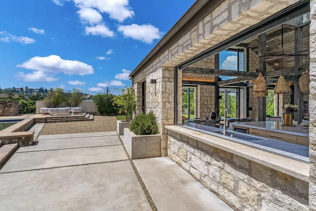 Modern outdoor patio with a stone building, a glass window, and a view of a backyard with a gravel pathway, seating area, and greenery under a blue sky with clouds.