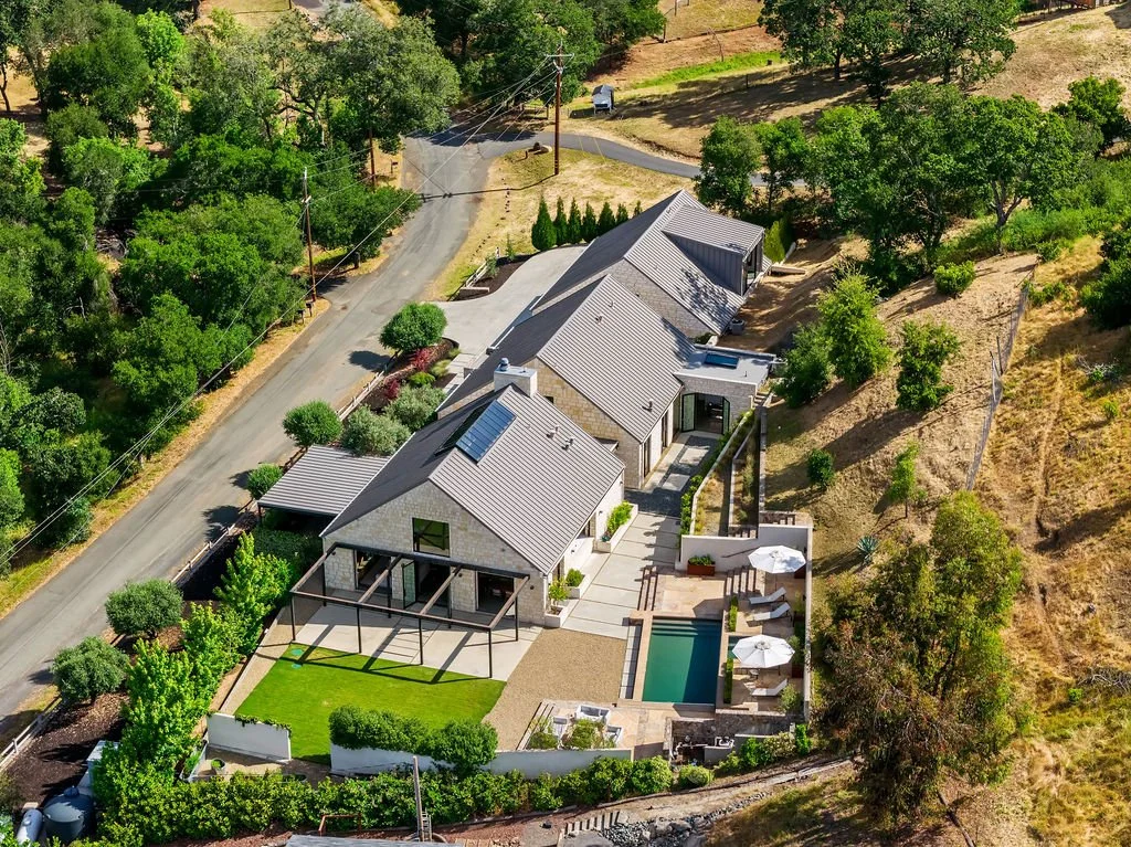 Aerial view of a modern house with a backyard pool, patio, and landscaped yard, surrounded by trees and located on a hillside.