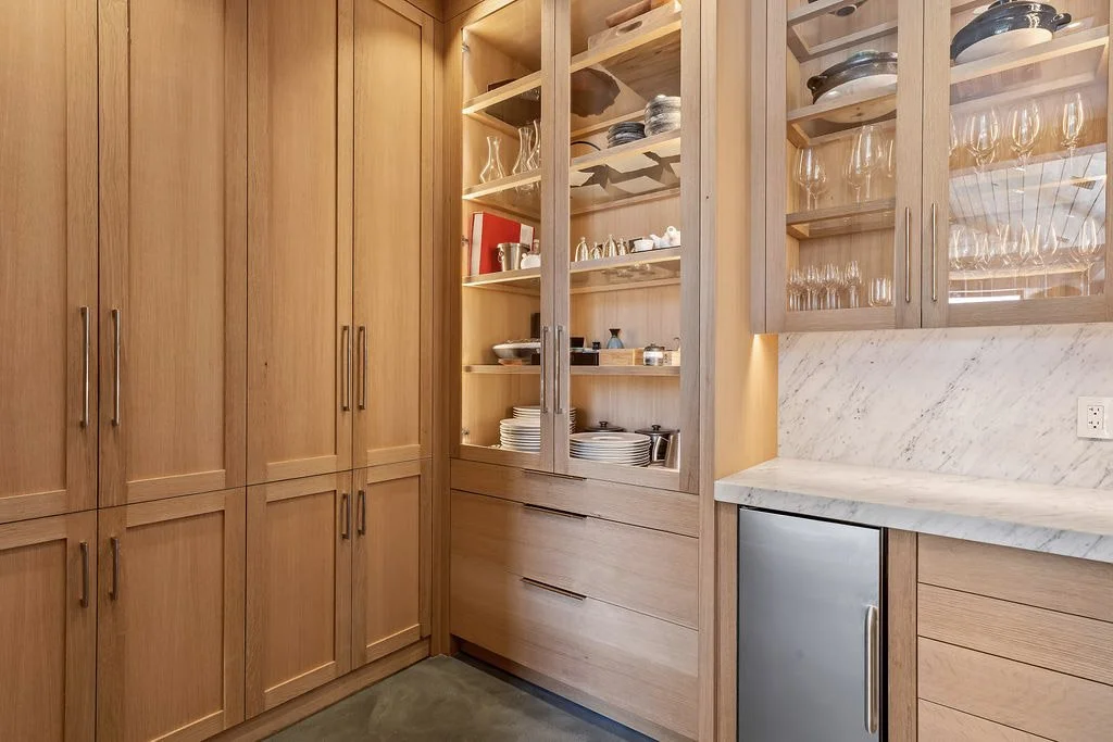 Wooden kitchen cabinets with glass doors filled with dishes and glassware, marble countertop, and a small mini fridge.