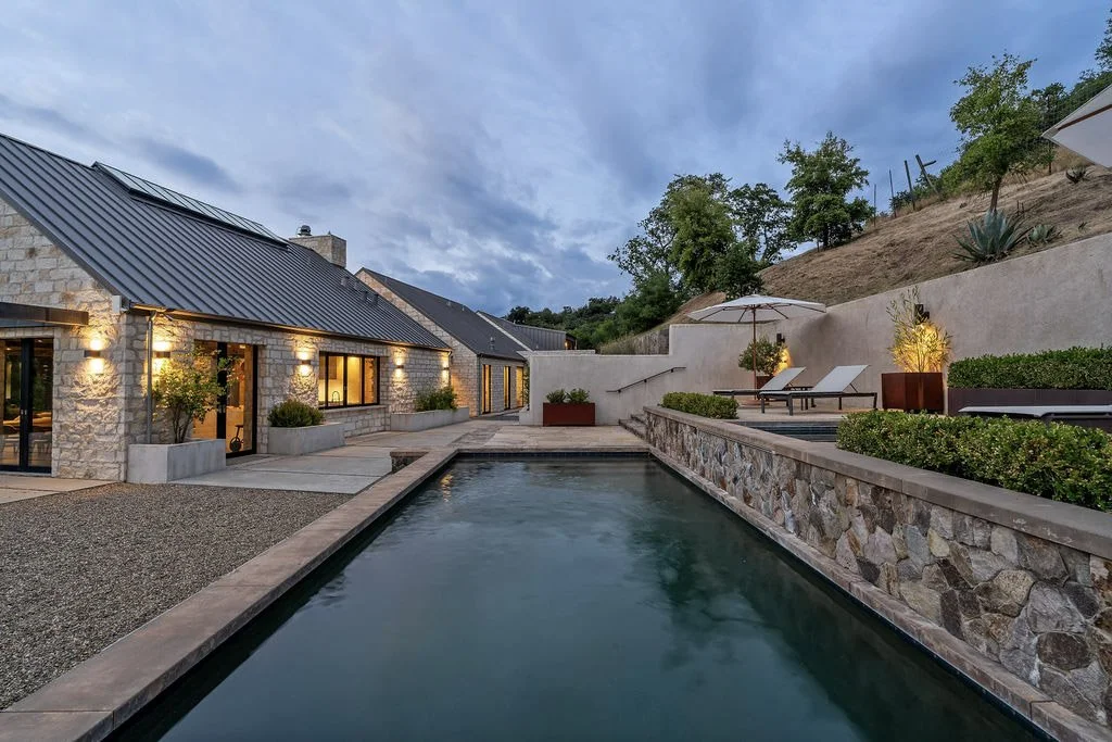 Modern backyard with a rectangular swimming pool, lounge chairs, umbrellas, potted plants, and a stone house with lights