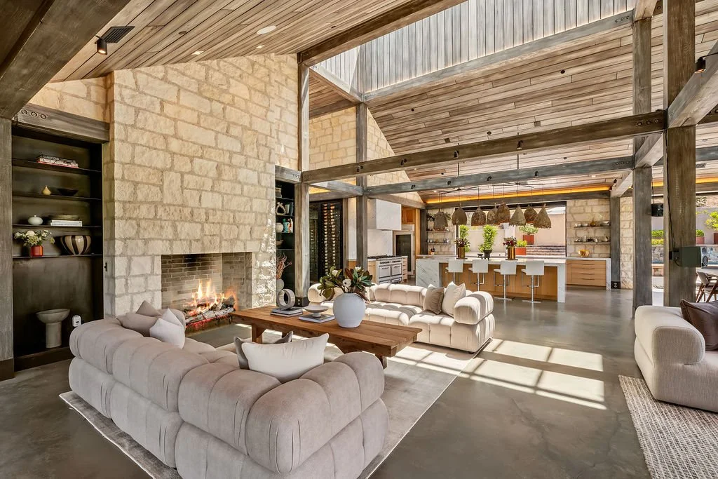 Spacious open-concept living room with stone fireplace, beige sectional sofa, wooden coffee table, and modern kitchen with bar stools and hanging pendant lights.