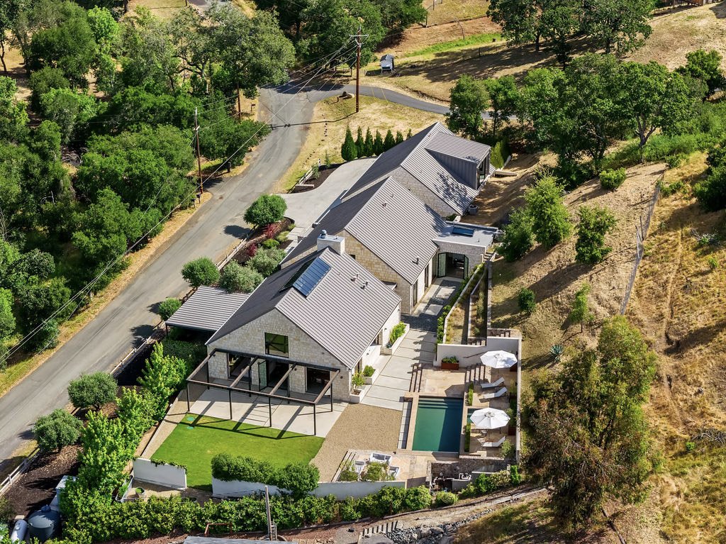 Aerial view of a modern house with a backyard featuring a swimming pool, patio area with umbrellas, and well-maintained lawn, surrounded by trees and natural landscape on a hillside.