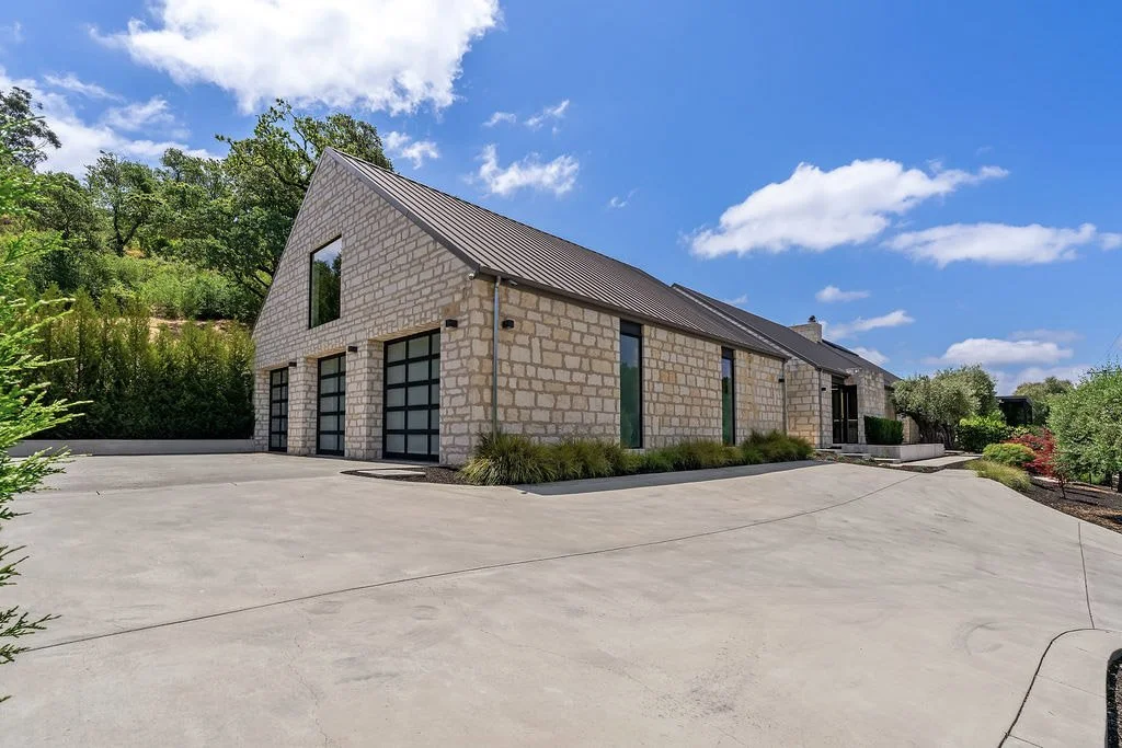 Modern house with stone exterior, metal roof, large ground-level garage doors, and tall windows, surrounded by greenery and trees under a partly cloudy sky.