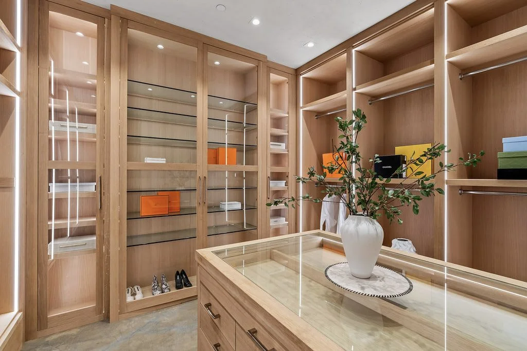 A walk-in closet with wooden shelves and drawers, some containing colorful boxes and shoes, a glass-fronted cabinet, and a white vase with green branches on a wooden island