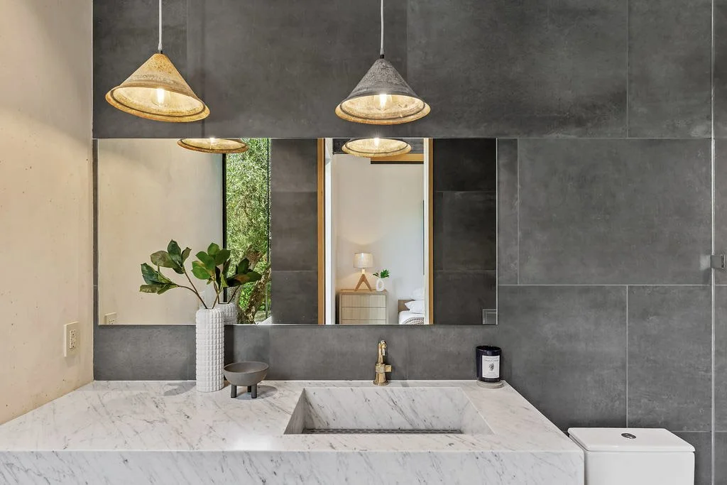 Modern bathroom with marble countertop, large mirror, grey tiled wall, green houseplant in a white textured vase, small bowl, candle, and gold faucet, reflected in a mirror showing a bedroom with a lamp and bed.