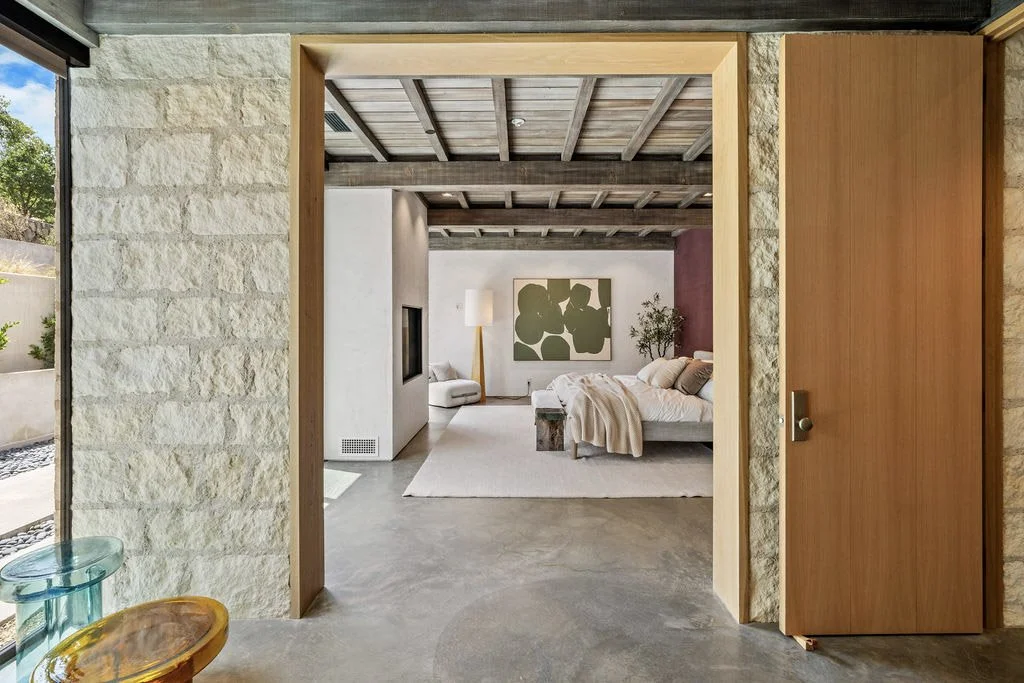 A view of a modern bedroom seen through an open doorway with stone and wood walls. The bedroom features a bed with neutral bedding, a large piece of abstract wall art, a lamp, a potted plant, and a cozy armchair.